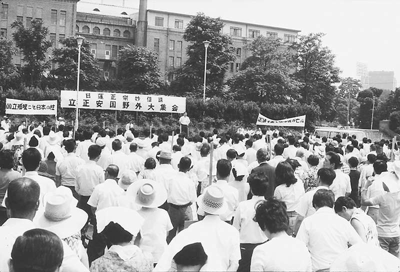 野外会場に多数の参加者が集まり、壇上の前に横断幕が掲げられている白黒写真