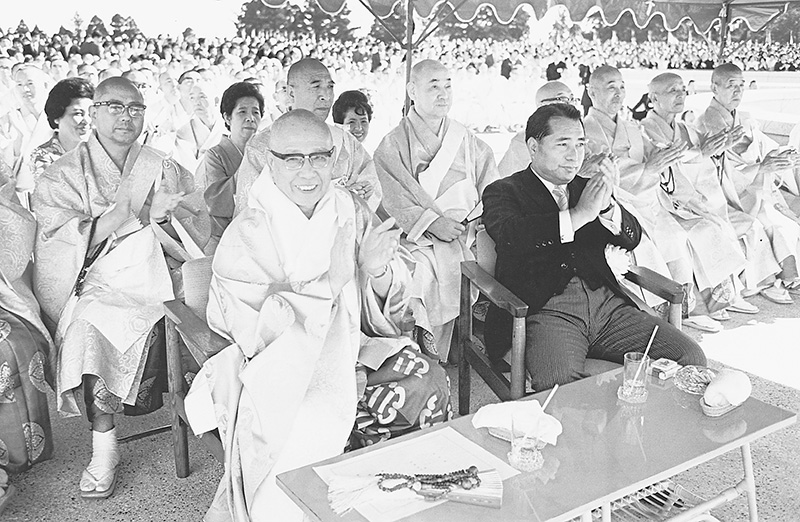 式典会場で法衣姿の僧侶やスーツ姿の男性らが椅子に並んで座り、後方に多数の参列者が見える白黒写真