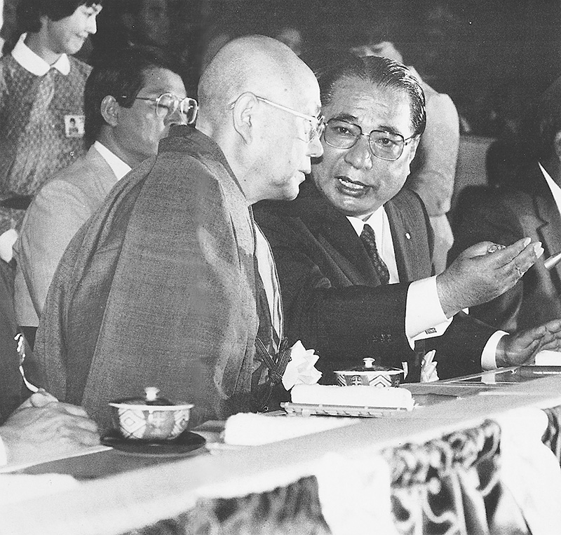 法衣姿の阿部日顕とスーツ姿の池田大作が、式典の席で隣同士に座り話している白黒写真