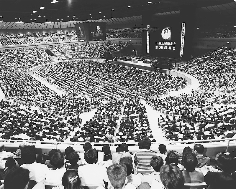横浜アリーナの会場いっぱいに多数の参加者が集まり、正面の壇上と大型スクリーンが見える白黒写真