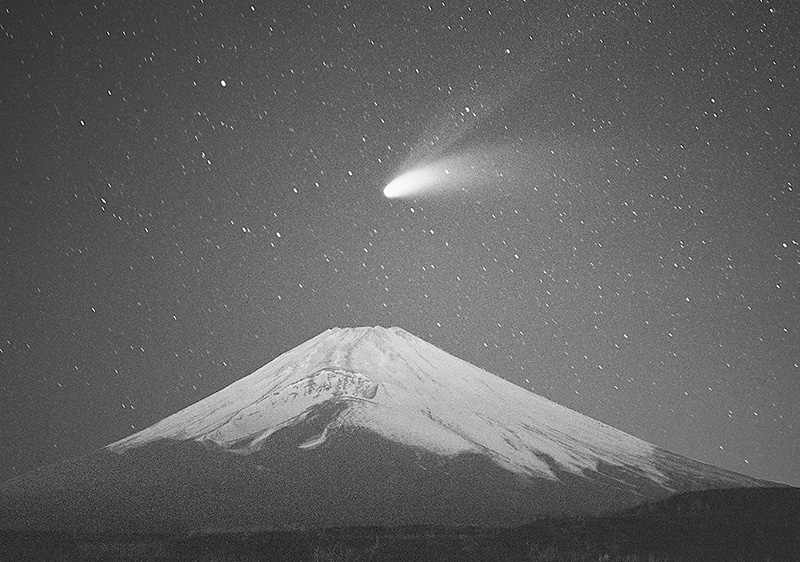 星空の下、富士山の上空に明るい尾を引く彗星が見える白黒写真